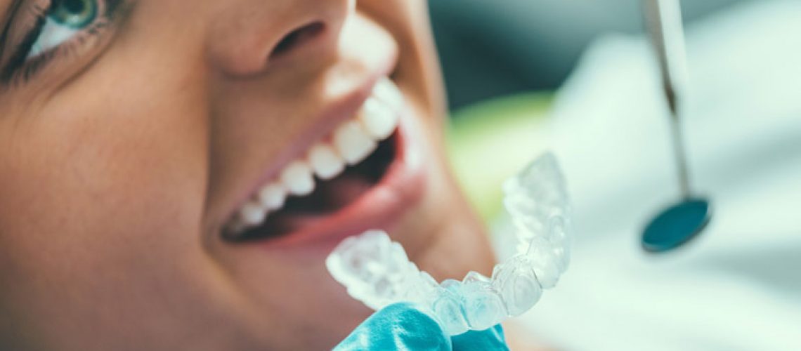 Close up photo of a doctor holding a clear aligner up to a female patient’s teeth.